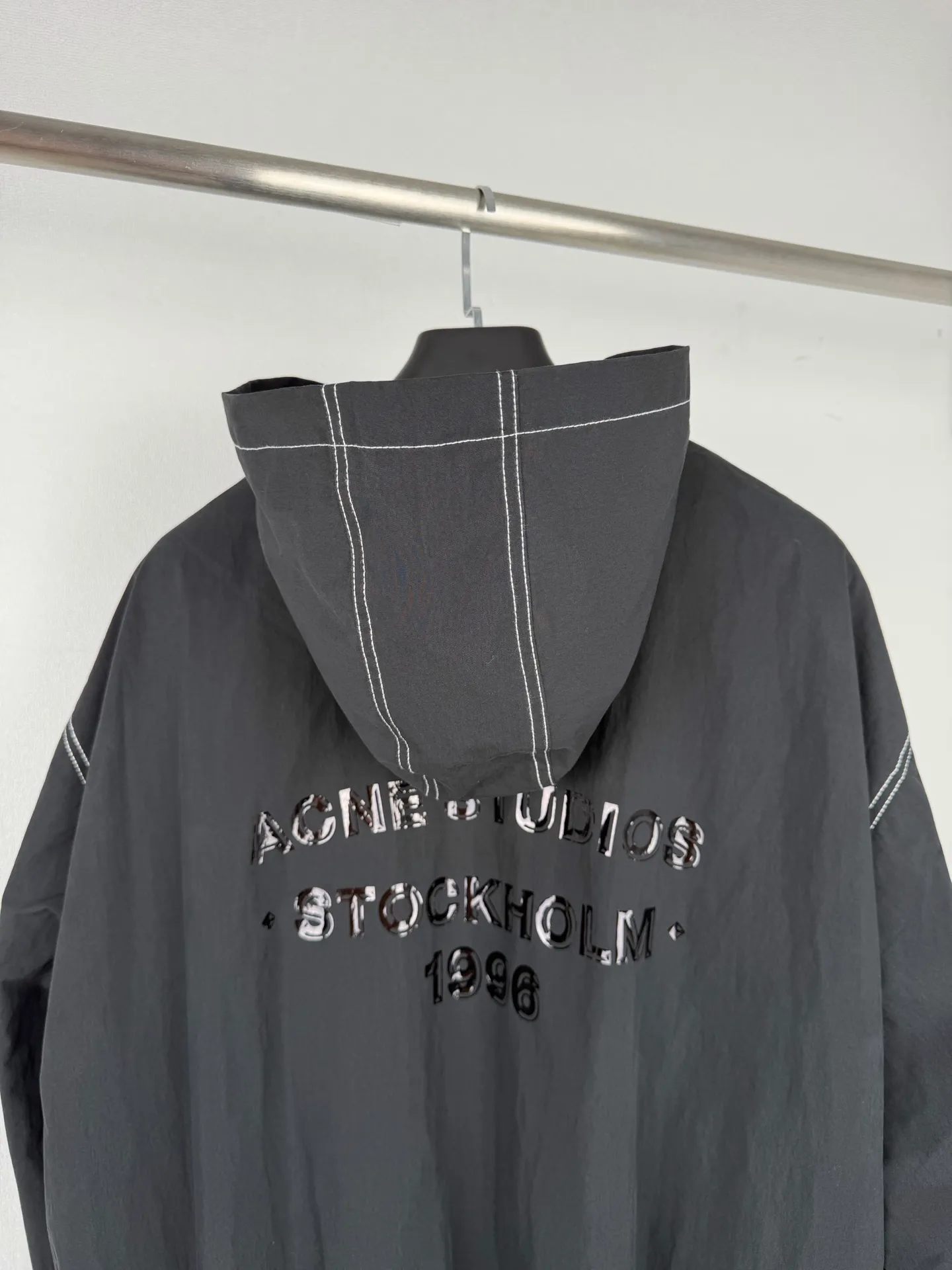ACNE Studios back letter topstitch hooded jacket is made of fabric processing, contrasting topstitching, printing alignment and combination of elastic components. The fabric is treated with a physical wrinkle process, which does not rely on chemical coatings. It uses mechanical wrinkles to create a disordered texture effect on the surface of the fabric. While the fabric has a relaxed look, it still retains the structural tension of the woven fabric. During the cutting stage, the deformation caused by the folds of the fabric needs to be fully considered to ensure a stable silhouette after the pieces are spliced. The entire product uses contrasting topstitching as the main structural decoration. The seams of the cap, shoulder seams, cuffs, and hem are all exposed with contrasting topstitching. There are arc turns in different parts of the cut pieces. When sewing, it is necessary to control the fabric feeding rhythm and maintain a uniform stitch density. The elastic area of ​​the hem is sewn with multiple layers of materials. The layers of fabric, elastic bands, and hemming pieces are overlapped. It is easy for the line to shift and the fabric to arch, which requires high precision in the lathe operation. The Retro character printing on the back is the visual core of the entire garment. Multiple groups of characters are arranged in layers, giving it a worn and broken texture. The printing must ensure that the layer is strong and washable, but cannot cover up the details of the incomplete retro characters. At the same time, the printing positioning needs to be aligned with the middle seam of the back of the garment to avoid tilting and offset of the printing. The cuffs and hem are both elastic pleated structures. When sewing, you need to control the density of the fabric pleats to ensure a uniform and natural arc after shrinkage. The hood is spliced ​​into pieces and relies on multiple open threads to separate and shape it, ensuring that the hat is natural and three-dimensional and will not collapse against the face when worn. After all the sewing processes are completed, the garment needs to be ironed and finished to unify the wrinkles of the entire fabric and remove excess creases caused by the production process, finally presenting the complete Retro relaxed garment effect. Size: 46 48 50 52